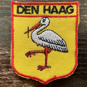 May include: A shield-shaped embroidered patch featuring a yellow background with a red border. The text DEN HAAG is stitched in black at the top. A white stork with black wing details, a red beak, and red legs stands in the center of the patch.