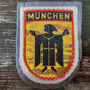 May include: A rectangular embroidered patch with a grey felt border. The center features a yellow shield with a black monk figure, known as the Münchner Kindl, and the text MÜNCHEN in black letters on a yellow background at the top.