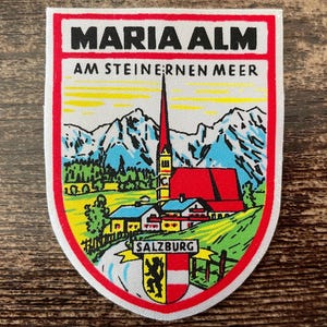 Könnte beinhalten: Ein schildförmiger Aufnäher mit einer Landschaft von Maria Alm am Steinernen Meer, inklusive Kirche, Bergen und grünen Wiesen. Der Text lautet MARIA ALM, AM STEINERNEN MEER und SALZBURG über einem Wappen mit einem schwarzen Löwen und rot-weißen Streifen.
