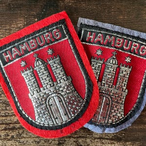 Könnte beinhalten: Zwei schildförmige, bestickte Aufnäher mit dem Hamburger Wappen. Jeder Aufnäher zeigt eine silberne Burg mit drei Türmen auf rotem Grund, darüber der Schriftzug HAMBURG in silbernem Garn, eingefasst mit schwarzer Naht auf Filzstoff.
