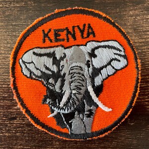 May include: Embroidered patch featuring an elephant with gray and black details, set against an orange background. The word "KENYA" is stitched in black above the elephant. The patch has a black border.