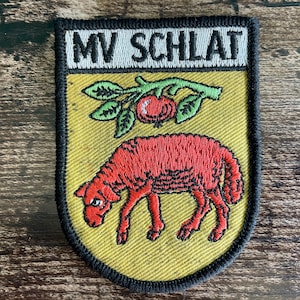 Könnte beinhalten: Ein schildförmiger, bestickter Aufnäher mit schwarzem Rand. Der obere Bereich zeigt den Text MV SCHLAT in Schwarz auf weißem Grund. Der untere gelbe Teil zeigt ein rotes Schaf unter einem grünen Zweig mit einer einzelnen roten Frucht.