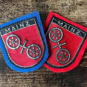 May include: Two embroidered patches with the word "MAINZ" at the top. The patches are shield-shaped with a red center and a silver emblem of two wheels. One patch has a blue border, the other a red border.