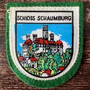 Könnte beinhalten: Ein schildförmiger Aufnäher mit grünem Filzrand und weißer Zickzacknaht. In der Mitte befindet sich eine farbenfrohe Illustration von Schloss Schaumburg mit roten Dächern, grauen Steinmauern und grünen Bäumen vor einem hellblauen Himmel.