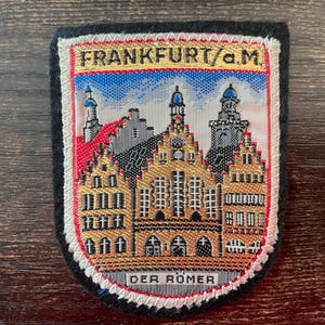 Könnte beinhalten: Ein rechteckiger gestickter Aufnäher mit einer gewebten Szene des Frankfurter Rathauses, bekannt als Römer. Der Text FRANKFURT/a.M. steht oben und DER ROMER unten. Der Aufnäher hat einen schwarzen Rand und eine weiße gestickte Kante.