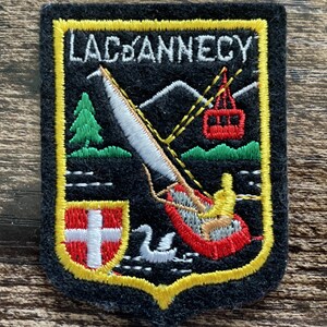 Könnte beinhalten: Ein schwarzer bestickter Aufnäher mit gelbem Rand und dem Text LAC D'ANNECY. Das Design zeigt eine rote Seilbahn, einen Windsurfer, einen grünen Tannenbaum, einen weißen Schwan und ein Wappen mit weißem Kreuz auf rotem Grund.