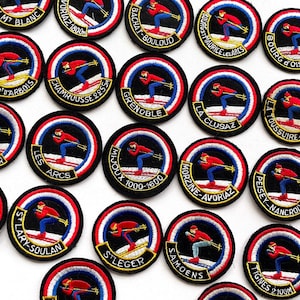May include: A collection of circular embroidered patches featuring a skier in a red jacket and blue trousers. Each patch displays a unique French ski resort name and elevation in white text against a black background with a red, white, and blue border.