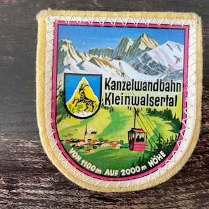 May include: A vintage patch featuring a mountain scene with a cable car, a village, and the text "Kanzelwandbahn Kleinwalsertal". The patch has a yellow border with white stitching and a pink inner border. The text indicates an elevation of 1100m to 2000m.