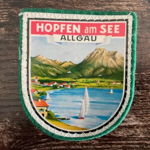 Puede incluir: Un parche de recuerdo en forma de escudo con una colorida ilustración de un lago, veleros, un pequeño pueblo y montañas. El texto "HOPFEN am SEE" y "ALLGÄU" está impreso en blanco sobre una bandera roja y blanca en la parte superior.
