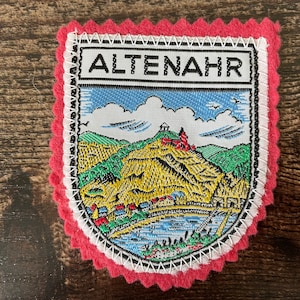 Op de afbeelding: Een rechthoekige geborduurde patch met een roze geschulpte vilten rand. De patch toont een landschapsillustratie van een stad, een rivier en een heuvel met een kasteel, bekroond met de tekst ALTENAHR in zwarte letters op een witte achtergrond.