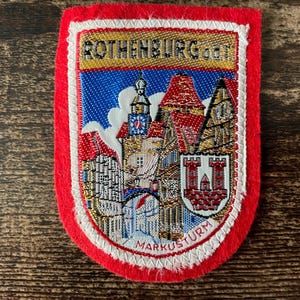 May include: Embroidered patch with a red border, depicting a cityscape with buildings and a tower. The patch features the text "ROTHENBURG" and "MARKUSTURM". The design is set against a blue and white sky, with a white and gold trim.
