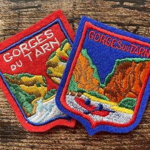 May include: Two shield-shaped embroidered patches featuring the text GORGES DU TARN. One patch has a red border and background with a river and canyon landscape. The other has a blue border and red inner frame with a canyon and a red raft.