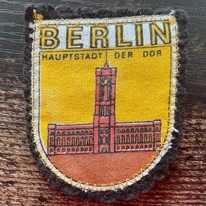 May include: A fabric patch featuring a yellow background with a red illustration of the Rotes Rathaus building in Berlin. The text BERLIN and HAUPTSTADT DER DDR are printed in black. The patch has a black scalloped border with yellow stitching.