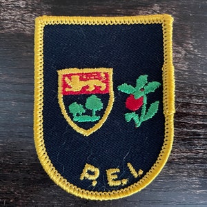 May include: A dark blue embroidered patch with a yellow stitched border. The patch features a shield with a gold lion over green trees, a red berry with green leaves, and the yellow embroidered letters P.E.I. at the bottom of the fabric.