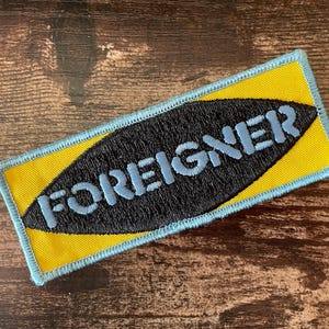 May include: A rectangular patch with the word "FOREIGNER" embroidered in light blue on a black oval. The oval is set against a yellow background, framed by a light blue border.