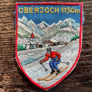 Könnte beinhalten: Ein schildförmiger gestickter Aufnäher mit einer Berglandschaft und dem Text OBERJOCH 1150m oben. Die Szene zeigt einen Skifahrer in roter Jacke und blauer Hose auf einem verschneiten Hang, mit einem kleinen Dorf, einer Kirche und Berggipfeln.