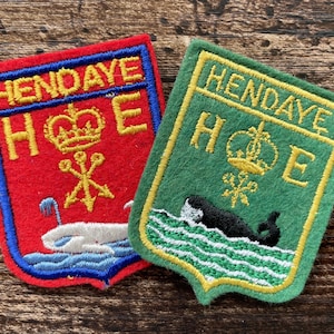 May include: Two shield-shaped embroidered patches featuring the text HENDAYE and H E with a crown and trident symbol. The red patch displays a white whale in blue waves, while the green patch shows a black whale in green and white waves.