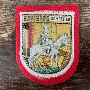 May include: A red, shield-shaped patch with the text "BAMBERG DOMREITER" at the top. The patch features a detailed image of a person riding a white horse. The image is surrounded by a white border and a red felt edge.