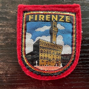 May include: A rectangular embroidered patch with a thick red felt border. The center features a woven scene of the Palazzo Vecchio in Florence, Italy, against a blue sky with white clouds. The word FIRENZE is stitched in gold at the top.