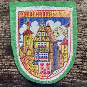 Könnte beinhalten: Ein schildförmiger, bestickter Aufnäher mit grünem Filzrand, der die Architektur von Rothenburg ob der Tauber zeigt. Der Text ROTHENBURG o.d. Tauber ist oben über einem zentralen Fachwerkhaus, einem Turm und einem roten Burgwappen unten vor einem blauen Himmel mit weißen Wolken eingestickt.