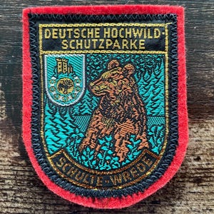 May include: A rectangular embroidered patch with a bright red felt border. The centre features a brown bear illustration against a teal forest background, a small crest with a castle, and the text DEUTSCHE HOCHWILD-SCHUTZPARKE and SCHULTE-WREDE.