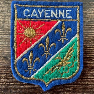 Könnte beinhalten: Ein schildförmiger, bestickter Aufnäher mit blauem Rand und dem Wort CAYENNE in weißem Garn. Der Schild ist in drei Bereiche unterteilt, die eine goldene Sonne auf Rot, drei goldene Lilien auf Blau und einen goldenen Vogel auf Grün zeigen.