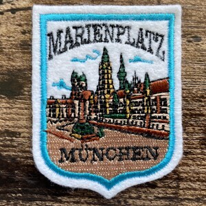 May include: A shield-shaped embroidered patch featuring a detailed depiction of the Marienplatz in München. The design includes black text, a central tower, and architectural elements in brown, green, and white, all bordered by a bright blue trim.
