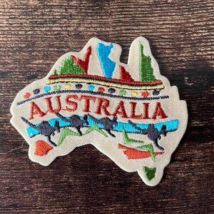 May include: Embroidered patch in the shape of Australia, featuring the word "AUSTRALIA" in red, kangaroos, and colorful landscape details. The patch is beige with a brown wooden background.