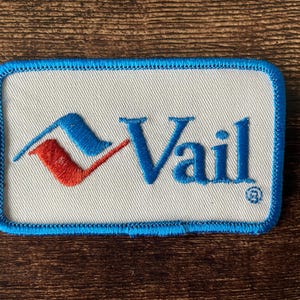 Op de afbeelding: Geborduurde patch met het woord "Vail" in blauw, met een gestileerd rood en blauw ontwerp. De patch heeft een witte achtergrond en een blauwe rand. De patch is rechthoekig en lijkt nieuw.