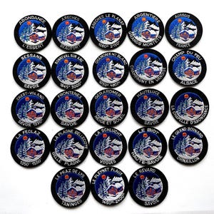 May include: A collection of embroidered patches with circular designs. Each patch features a winter landscape with a cabin, trees, and text indicating a location. The patches are black with blue and white details, and the text is in white.