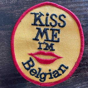 Könnte beinhalten: Ein kreisförmiger bestickter Aufnäher mit gelbem Hintergrund und rotem Rand. Der Aufnäher zeigt den schwarzen Schriftzug "KISS ME I'M Belgian" mit einem roten, gestickten Lippenpaar, das zwischen den Wörtern "I'M" und "Belgian" platziert ist.