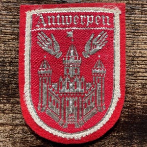 Può includere: Una toppa ricamata rossa raffigurante un castello argentato, due mani mozzate e una piccola bandiera in cima. La parola Antwerpen è cucita con filo d'argento nella parte superiore. La toppa ha un bordo bianco e una forma a scudo.