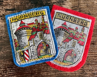 May include: Two rectangular felt patches featuring embroidered cityscapes of Nurnberg. One patch has a blue felt border, and the other has a red felt border. Both patches display a detailed illustration of a castle gate, towers, and the text NURNBERG.