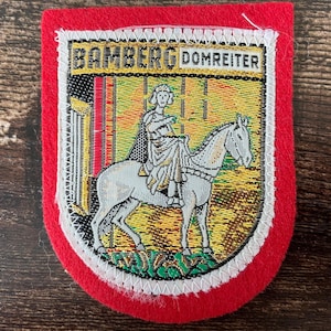 Peut inclure: Un écusson rectangulaire en feutrine rouge présentant une scène brodée d'un cavalier sur un cheval blanc. La bannière supérieure affiche le texte BAMBERG DOMREITER en lettres noires sur fond jaune. L'image centrale montre une silhouette à cheval.