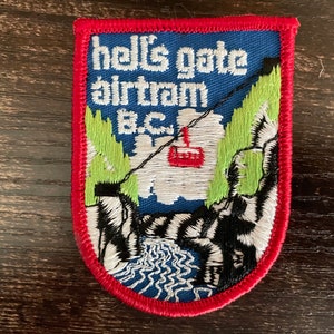 Op de afbeelding: Een rechthoekige geborduurde patch met een rode rand, voorzien van een blauwe lucht, groene bomen en witte bergen. Een rode kabelbaanwagen hangt aan een zwarte kabel. De tekst "hell's gate airtram B.C." staat in witte letters boven een rivier.