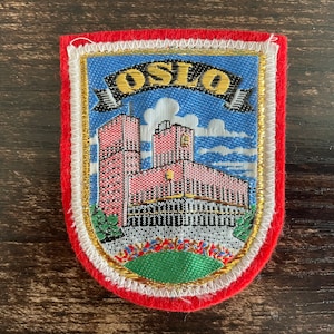 May include: Embroidered patch featuring the city name "OSLO" above a depiction of buildings and a blue sky with clouds. The patch has a red border, white and gold trim, and a green base.