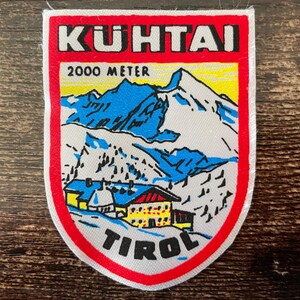 Könnte beinhalten: Ein schildförmiger Aufnäher mit einer Berglandschaft, schneebedeckten Gipfeln, einem gelben Gebäude und dem Text KUHTAI, 2000 METER sowie TIROL. Das Motiv ist von einem dicken roten Rand auf weißem Grund mit schwarzer Schrift umgeben.