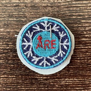 Peut inclure: Un écusson brodé circulaire présentant un anneau extérieur bleu foncé avec un motif de flocon de neige blanc. Le centre est un cercle bleu clair avec le texte rouge ÅRE brodé au milieu. Une petite épingle en métal argenté est fixée.