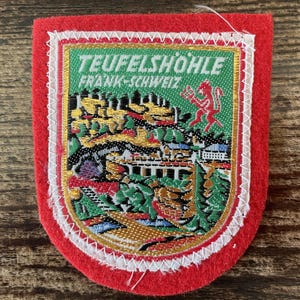 May include: A red felt patch featuring a shield-shaped embroidered emblem with the text TEUFELSHÖHLE FRANK-SCHWEIZ. The design depicts a landscape with trees, a bridge, and a red devil figure on a green background, bordered by white stitching.