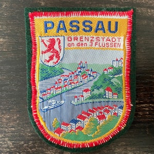 May include: Embroidered patch featuring the city of Passau, Germany. The patch has a shield shape with a green border, red and yellow stitching, and a detailed image of the city with text that reads "PASSAU". A crest is on the left side.