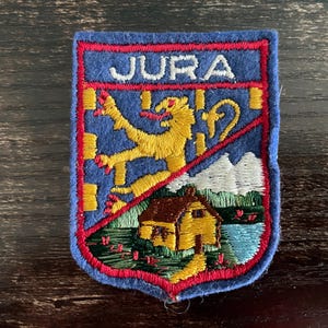 Puede incluir: Un parche bordado rectangular con borde azul y ribete rojo. La sección superior presenta la palabra JURA en letras blancas sobre un león amarillo en un fondo azul. La sección inferior representa una casa amarilla cerca de un lago y montañas.