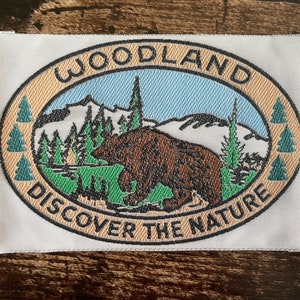 May include: Embroidered patch with an oval design featuring a brown bear, mountains, and trees. The text "WOODLAND DISCOVER THE NATURE" is arched around the image in a tan and black color scheme. The patch is on a white background.