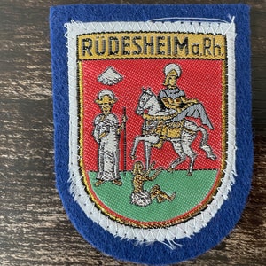 Op de afbeelding: Een blauwe vilten patch met een geborduurd wapen. Het wapen toont figuren op een paard en staand, met de tekst "RÜDESHEIM a.Rh." bovenaan. De patch heeft een witte rand en ligt op een donkere houten ondergrond.