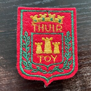 Op de afbeelding: Een rode viltpatch in de vorm van een schild met een groene geborduurde rand. Het heeft een gouden geborduurde kroon aan de bovenkant, de tekst THUIR boven drie gouden torens en de tekst TOY onder de torens, omringd door groene lauriertakken.