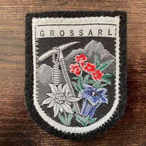 Könnte beinhalten: Gestickter Aufnäher mit dem Wort "GROSSARL" oben. Der Aufnäher zeigt eine Berglandschaft mit einem Eispickel, roten, blauen und weißen Blumen und einem weißen Rand auf schwarzem Hintergrund.