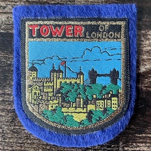 Op de afbeelding: Een schildvormige geborduurde patch met een blauwe vilten rand. Het midden toont een gedetailleerde scène van de Tower of London in geel en zwart, tegen een helderblauwe lucht met wolken. De tekst TOWER OF LONDON is in rood en zwart gestikt.