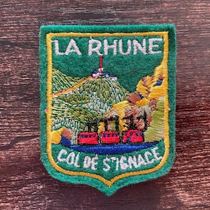 Pode incluir: Um emblema bordado verde apresentando uma paisagem de montanha com um trem vermelho na base. O texto LA RHUNE está bordado em branco no topo e o texto COL DE STIGNACE em branco na parte inferior, tudo contornado por uma linha amarela.