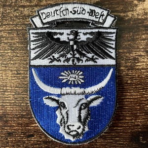 May include: A shield-shaped embroidered patch featuring a black eagle on a white background above a blue field with a white ox head and a small sun symbol. The top banner displays the black text Deutsch-Süd-West on a white background.