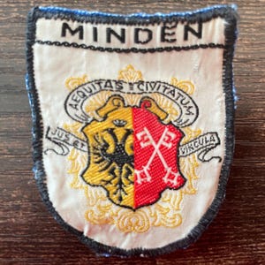 Puede incluir: Un parche bordado en forma de escudo con fondo blanco y borde negro. El texto "MINDEN" aparece en la parte superior. Un escudo central muestra un águila negra sobre campo dorado y dos llaves blancas sobre campo rojo, rodeado de texto en latín.