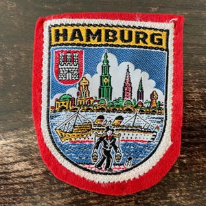 May include: Embroidered patch with a red border and the word "HAMBURG" at the top. The patch features a detailed illustration of Hamburg's skyline, ships, and a figure carrying buckets. The background is blue with white stitching.